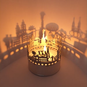 Puede incluir: Un portavelas de metal dorado con un diseño de ciudad. El portavelas está encendido y proyecta sombras en la pared detrás de él. El diseño de la ciudad incluye edificios, una torre y una noria. El texto "moominmamma" es visible en el portavelas.