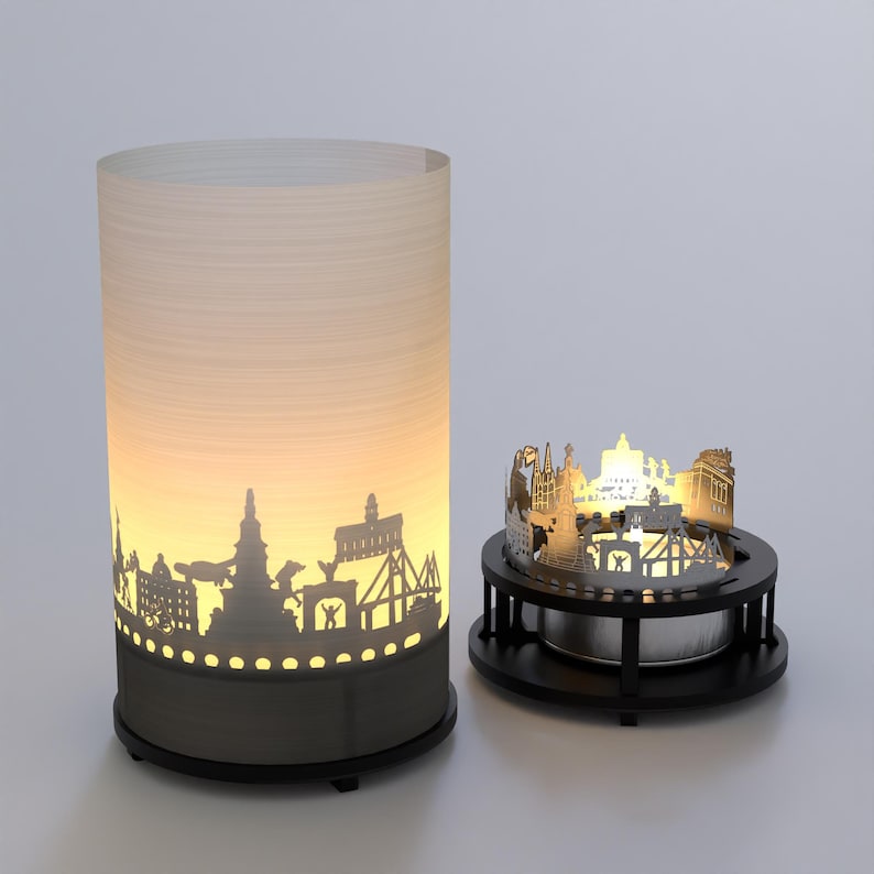 May include: Two candle holders with a cityscape silhouette design. The larger holder is a cylinder with a white frosted glass exterior and a black base. The smaller holder is a round metal base with a cityscape silhouette design and a small candle holder in the center.