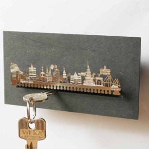 May include: A silver metal key holder shaped like the Savannah, Georgia skyline. The key holder is mounted on a gray background. A key is hanging from the holder.