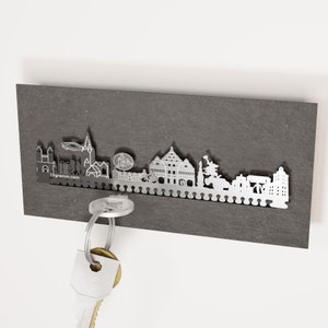 May include: A silver metal key holder shaped like the city skyline. The key holder is mounted on a dark gray slate background. A key is hanging from the key holder.