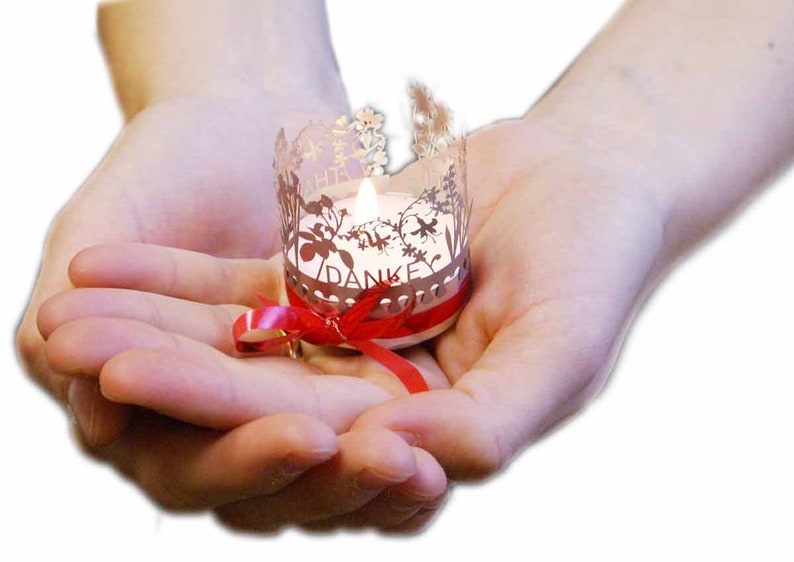 May include: A small, white candle with a red ribbon is held in cupped hands. The candle is inside a silver holder with a delicate floral design and the word "DANKE" inscribed on it.
