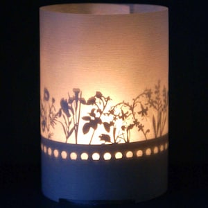 May include: A white paper candle holder with a silhouette of a garden scene cut out. The candle holder has a row of small holes around the bottom edge.