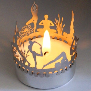 May include: Silver candle holder with a cutout design of yoga poses and a lit tea light candle inside.