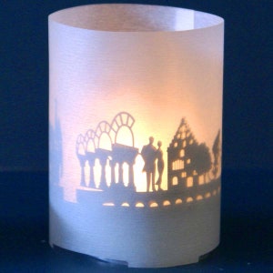 Puede incluir: Una linterna de papel blanca con una silueta de un horizonte de ciudad, incluyendo edificios, árboles y dos personas. La linterna está iluminada desde adentro, proyectando un brillo cálido.
