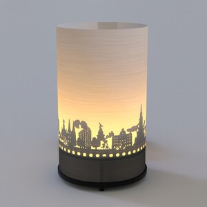 May include: A white cylindrical lamp with a cityscape silhouette cut out. The lamp is lit from within, casting a warm glow through the cutouts. The base of the lamp is black.