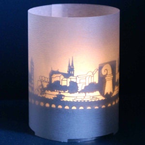 Puede incluir: Una linterna de papel blanco con un diseño de silueta de ciudad. La linterna está iluminada desde adentro, proyectando un brillo cálido a través de los recortes.