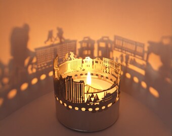May include: A gold metal candle holder with a cityscape design. The candle holder is lit and casting shadows on the wall behind it.