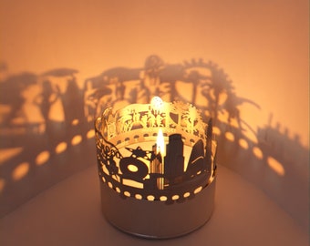 May include: A white metal candle holder with a cityscape design. The candle holder is lit and casting a shadow on the wall behind it.