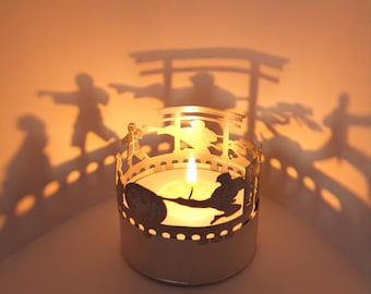 May include: A silver candle holder with a cut-out design of people and a torii gate. The candle holder is lit with a tea light candle, casting shadows on the wall.