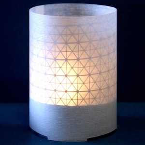 Peut inclure: Un bougeoir cylindrique blanc avec un motif géométrique. Le bougeoir est allumé et la lumière brille à travers le motif.