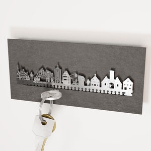 May include: A silver metal key holder with a city skyline silhouette design, mounted on a dark grey rectangular board. The key holder has small hooks for keys. The website address "13gramm.com" is visible. Keys are hanging from the holder.