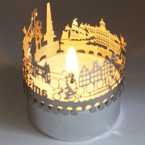 May include: A silver candle holder with a cutout design of a city skyline. The candle holder is lit with a candle inside.
