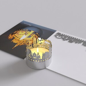 May include: A silver candle holder with a cutout design of the Vienna skyline. The candle holder is sitting on a white surface with a postcard featuring a similar design in the background.
