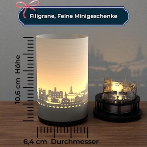 Puede incluir: Un portavelas cilíndrico blanco con un diseño de silueta de ciudad. El portavelas tiene 10,6 cm de alto y 6,4 cm de diámetro. El portavelas está encendido y proyecta un brillo cálido.
