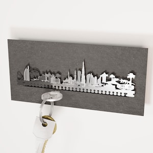 Könnte beinhalten: Ein silberner Schlüsselhalter aus Metall in Form der Skyline von Dubai. Der Halter ist an einer dunkelgrauen Wand montiert und hat einen Schlüsselring daran.