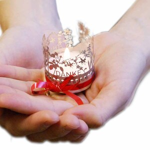Puede incluir: Una pequeña vela blanca con una cinta roja se sostiene en manos ahuecadas. La vela está dentro de un portavelas plateado con un delicado diseño floral y la palabra "DANKE" inscrita en él.
