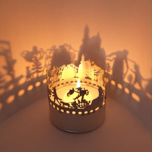 May include: A white metal candle holder with a cutout design of a fairytale scene. The candle holder is lit and casting shadows on the wall behind it.