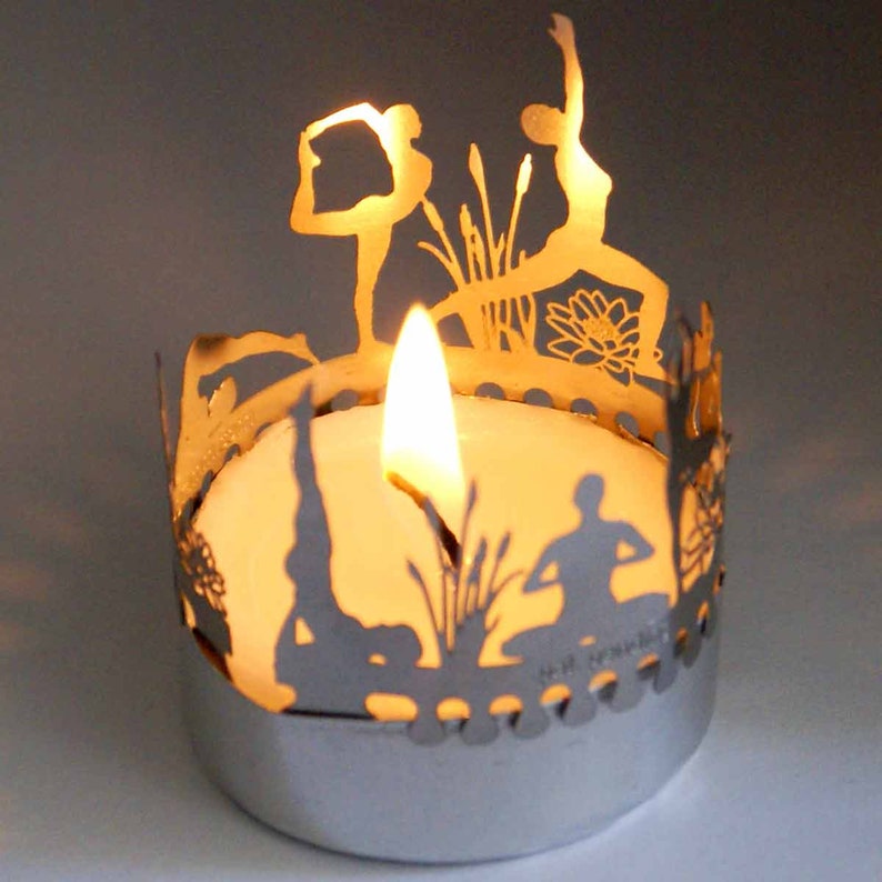 May include: Silver candle holder with a cutout design of yoga poses and lotus flowers. A white tea light candle is burning inside the holder.