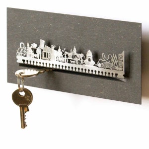 Könnte beinhalten: Ein silberner Schlüsselhalter aus Metall in Form der Skyline von Leipzig. Der Halter hat einen Schlitz für Schlüssel und das Wort "LEIPZIG" ist seitlich aufgedruckt.