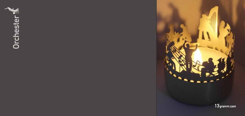 Puede incluir: Un portavelas de plata con una silueta de una orquesta tocando m&uacute;sica. El portavelas est&aacute; iluminado con una peque&ntilde;a vela de t&eacute;.