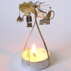 May include: A silver metal candle holder with a rotating carousel featuring cut-out silhouettes of buildings and figures. The carousel is labeled "Weimar" and has a lit tea light candle in the center.