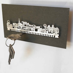 May include: A silver metal key holder shaped like the Durham, England skyline. The holder is mounted on a dark brown background. Two keys are hanging from the holder.