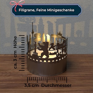 K&ouml;nnte beinhalten: Ein Kerzenhalter aus Metall mit einer Silhouette der Berliner Skyline, einschlie&szlig;lich des Brandenburger Tors, des Reichstagsgeb&auml;udes und des Berliner B&auml;ren. Der Kerzenhalter hat einen Durchmesser von 3,5 cm und eine H&ouml;he von 3 cm.