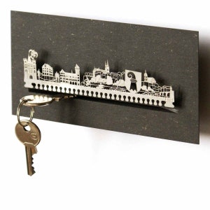 Puede incluir: Un porta llaves de metal plateado con forma de horizonte de una ciudad. El porta llaves está montado sobre un fondo gris oscuro. Una llave cuelga del porta llaves.