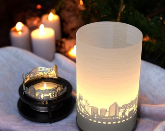 May include: Two candle holders with a cityscape design. The larger holder is white with a frosted finish and the smaller holder is silver with a cityscape silhouette. Both holders are lit with candles.