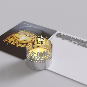 May include: A silver candle holder with a cut-out design of a city skyline. The candle holder is lit with a yellow candle inside.