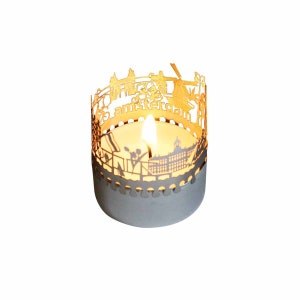 May include: A gold metal candle holder with a cut-out design of the Amsterdam skyline. The candle holder is lit with a small candle inside.