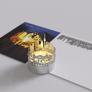 K&ouml;nnte beinhalten: Ein silberner Kerzenhalter mit einem ausgeschnittenen Design der Berliner Skyline. Der Kerzenhalter ist angez&uuml;ndet und die Flamme wirft einen warmen Schein auf die Umgebung. Der Text "Berlin" ist auf dem Kerzenhalter sichtbar.