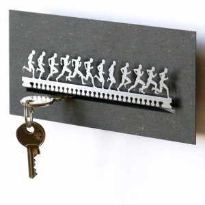 Può includere: Un portachiavi in ​​argento con una fila di figure che corrono ritagliate nel metallo. Una chiave è appesa al portachiavi.