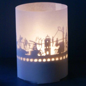 May include: A white paper lantern with a cityscape silhouette design. The silhouette includes buildings, windmills, and people. The lantern is lit from within, casting a warm glow.