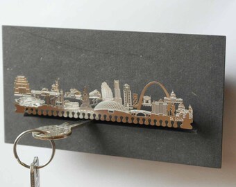 Könnte beinhalten: Ein silberner Schlüsselhalter aus Metall in Form der Skyline von St. Louis. Der Halter ist auf einem dunkelgrauen Schiefergrund montiert. Ein Schlüssel hängt am Halter.
