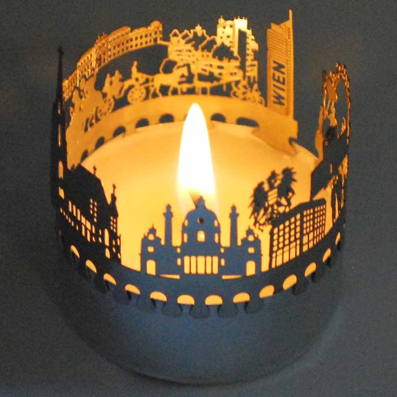 May include: A white metal candle holder with a cutout design of the Vienna skyline. The candle holder is lit with a candle inside, casting a warm glow.
