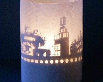 May include: A white paper candle holder with a cityscape silhouette cut out. The silhouette includes buildings, a car, and a boat.