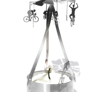 May include: A silver metal candle holder with a rotating carousel featuring silhouettes of cyclists. The carousel is suspended above a round candle holder with a white candle.
