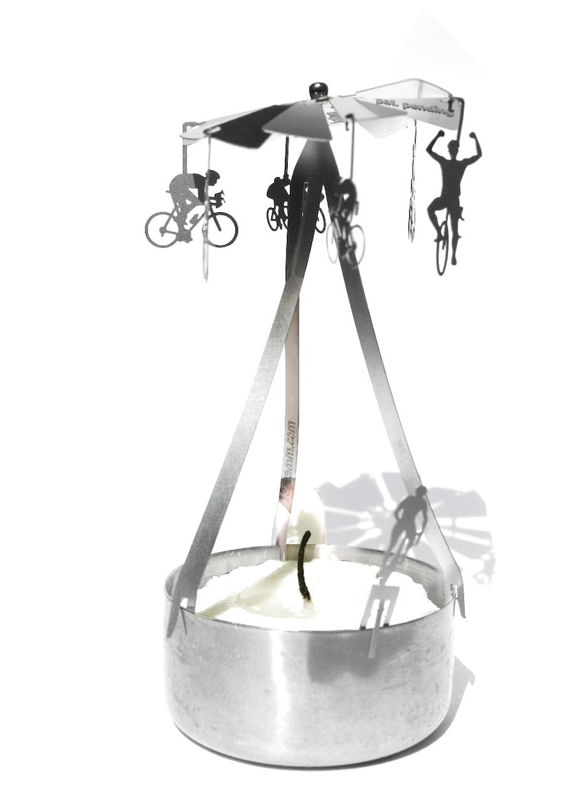May include: A silver metal candle holder with a rotating carousel featuring silhouettes of cyclists. The carousel is suspended above a round candle holder with a white candle.