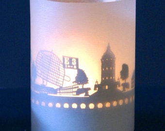 May include: A white paper candle holder with a silhouette of a city skyline. The skyline includes a Ferris wheel, a building with the text "E3" on a sign, and a tall tower. The candle holder has a decorative border of small circles around the bottom.