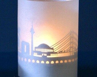 May include: White paper candle holder with a silhouette of a city skyline, including a bridge, a building with a dome, and a tall tower. The candle holder is lit, casting a warm glow.