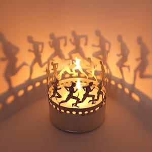 Può includere: Un portacandele in metallo bianco con un design di figure che corrono. Il portacandele è acceso e proietta ombre delle figure sul muro.