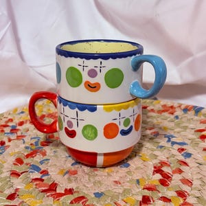 Puede incluir: Dos tazas de cerámica apiladas con un diseño caprichoso de cara de payaso. Las tazas son blancas con lunares de colores, detalles en forma de cruz y una cara sonriente. Una taza tiene un asa azul, la otra roja. Los bordes son festoneados y los interiores son amarillos.