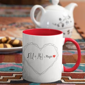 Puede incluir: Taza de cerámica blanca con interior y asa rojos. La taza presenta un diseño de corazón geométrico con el texto "S(U + Me) = Magia" y un pequeño corazón rojo. Una tetera blanca y galletas están en el fondo.