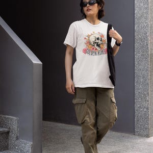 Puede incluir: Camiseta blanca con un diseño de calavera rodeada de hojas de otoño y el texto "GOLDEN ASHES". La persona lleva pantalones cargo verde oliva y gafas de sol negras. Una bolsa de mano negra está sobre el hombro.