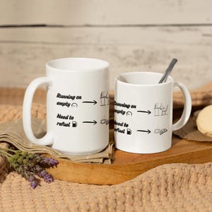 Puede incluir: Dos tazas de cerámica blanca con texto e ilustraciones en negro. El texto dice "Running on empty" y "Need to refuel". Una taza es más grande que la otra. Una cuchara descansa en la taza más pequeña.