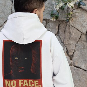 Op de afbeelding: Witte hoodie met een afbeelding op de achterkant. De afbeelding toont een bivakmuts-gezicht in rood en zwart, omlijnd door een rode rechthoek. Gouden tekst eronder luidt "NO FACE, NO MERCY".