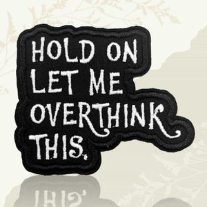 May include: Black embroidered patch with white text reading "HOLD ON LET ME OVERTHINK THIS." The patch has a distinctive, irregular shape with a black border. The text is in a bold, sans-serif font.