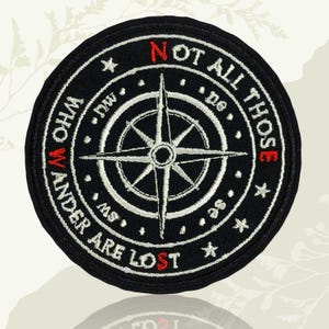 May include: A round, black embroidered patch featuring a white compass rose design. The patch includes the text "NOT ALL THOSE WHO WANDER ARE LOST" in white and red thread. Small stars and directional abbreviations are also present.