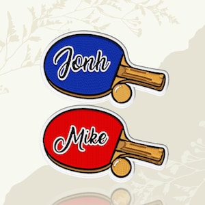 May include: Two embroidered ping pong paddle patches. One is blue with the name "Jonh" in white script, and the other is red with the name "Mike" in white script. Each patch includes a small orange ping pong ball.